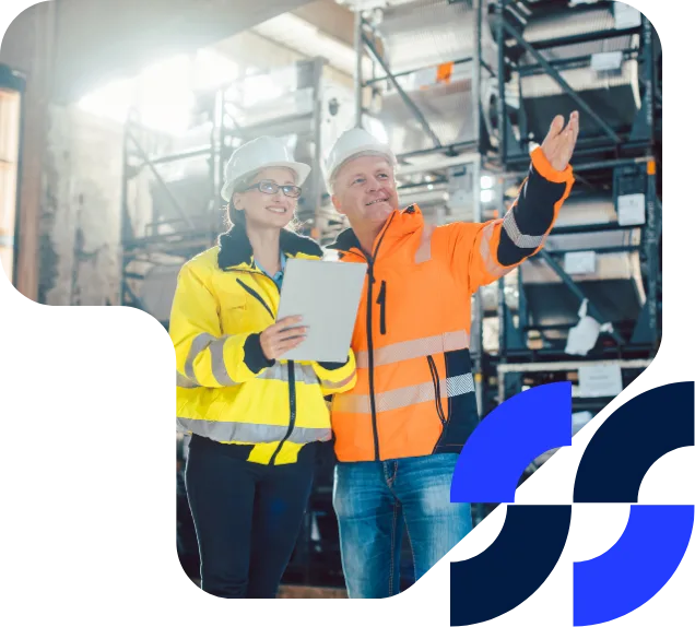 Two data centre engineers wearing high-visibility jackets and hard hats review plans in a warehouse, pointing toward a structure.